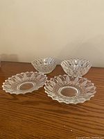 Two clear glass bowls and two clear glass plates with a floral petal motif around the edges shown side-by-side on a wooden surface.