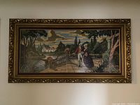 Framed needlepoint portrait depicting a man and woman in a garden setting with a water feature, in a large ornate gold frame