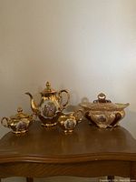 Full view of gold tea set on wood surface showing teapot, sugar bowl, creamer, and covered dish with ornate details and painted scenes.