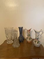 Five decorative vases and pitchers arranged on wooden surface against white wall.