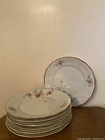 Stack of 7 round gold-rimmed porcelain plates with floral patterns, leaning against a single plate showing flower detail and gold rim.