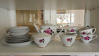 Photo showing several stacked plates, teapot, cups, sugar bowl, creamer, and saucers with pink rose pattern and gold trim inside a cabinet.