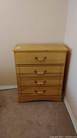 Full view of wood dresser with 4 drawers, light wood finish, metal handles, placed against wall on carpet.