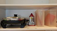 Black & Decker electric skillet with glass lid on shelf with metal box grater and two plastic containers with red lids.