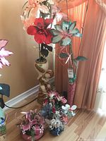 Photo showing various faux flower arrangements and decorative vases placed on a wooden floor against a beige wall near window drapes.