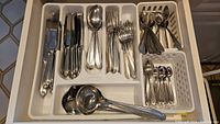 Top-down photo of a white utensil tray containing assorted flatware including knives, forks, spoons, ladle, and spatula.