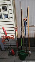 Photo showing the entire set of garden tools leaning against wall and door in sunlight: red Homelite trimmer with orange cord, long rake, spade, green watering can, garden fork, and hand cultivator.