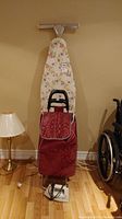 Full view showing the floral ironing board, red shopping cart leaning against it, and Norelco iron on floor near a lamp and wheelchair