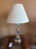 Two decorative table lamps with clear glass and shiny metal bases, white pleated shades, both standing on dark wooden surfaces, one lamp lit.
