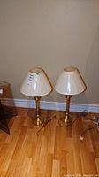 Photo of two brass finish table lamps with white fabric shades standing on a hardwood floor against a white wall. Both lamps plugged in with cords visible on floor.