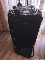 Black rolling suitcase standing upright with two silver folding carts on top and black purse in front.