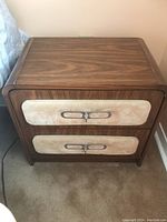 Front and side view of wood nightstand with cream fabric drawer fronts and metal handles.