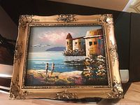 Photo of a larger framed painting showing a castle by the sea with two people on the shore, ornate gold frame visible.