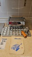 Front view of Titania pasta maker attached to ravioli tray with wooden handled crank and instruction booklet on counter.