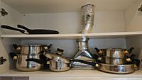 Photo showing an upper kitchen cabinet with multiple stainless steel pots, saucepans, and frying pans with black handles.