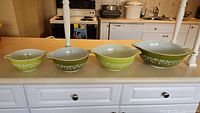 Four Pyrex mixing bowls in alternating lime and olive green colors with white floral designs and pouring spouts, displayed on a kitchen counter.