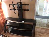 TV stand showing front left angle with two black tempered glass shelves, curved black legs, and central monitor mount brace with adjustable brackets.