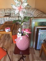 Pink ceramic vase with artificial flowers on a dark wooden table, surrounded by orange chairs and framed pictures in the background.