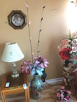 Full view showing vase sitting on the floor beside furniture and table with lamp, containing faux flowers with pink and purple leaves.