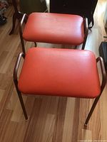 Front angled view of two orange vinyl stools with metal frames upright on wooden floor.