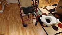Photo of the red and black Bios Diagnostics walker with front wheels and seat, standing on wooden floor near carpet.