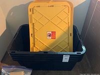 Four stacked black storage bins with one yellow lid resting inside.
