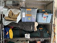 Metal shelving unit holding comforter, sleeping bag, green tent duffle, rolled blankets, and plastic storage totes labeled 'tools cooking' and 'stove'