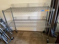 Full view of chrome two-tier shelving unit showing wire mesh shelves, vertical posts, guardrails, and caster wheels on carpeted floor near curtain.