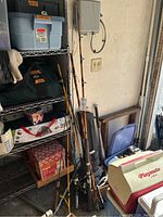 Photo showing multiple fishing rods with reels standing against a wall near shelves, tackle box visible on a shelf.