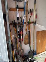 Full view of garden tools standing in corner including long handled shovels, rakes, hoes, pruning shears, broom, mop, and roll of garden tape.