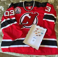 Koho New Jersey Devils 2003 Stanley Cup finals jersey laid out flat with 10 hockey pins in a box on top. Jersey is primarily red with black, white, and red striping and number 03 visible on sleeves.