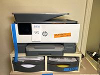 Front view of HP OfficeJet Pro 9018 printer on top of beige riser stand with four drawers containing labeled paper.
