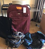 Large maroon Samsonite suitcase with front pocket and side handle, The North Face black backpack, black garment bag, two black and blue duffle bags, and gray travel pillow/headrest visible in a room.
