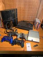 Two PlayStation 3 consoles (black fat model and slim model), four controllers, an optical drive for fat PS3, all displayed on a wooden surface.