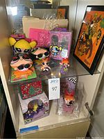 Display of plush dolls, mini figurines, bobble head, wardrobe set, light sculpture with documentation, and lunchbox arranged inside a cabinet.