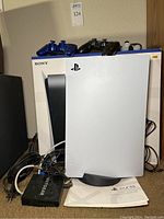 PS5 console standing in front of its box with three controllers on top, router and cables at base