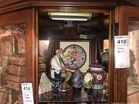 Photo of the cabinet shelf displaying the plate, porcelain figure, decorative eggs, and lidded jar