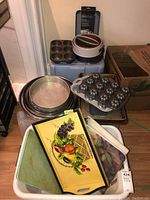 Wide view showing various baking pans, muffin tins, and colorful serving trays stacked on plastic bins and on the floor