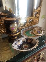Full view of the lidded jar, decorative bowl, platter, and wall hanging plate together on a table in front of a large ornate gold mirror