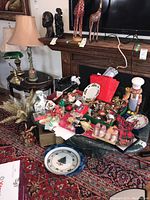 Overview of Christmas decorations, ornaments, plates, mugs on table next to fireplace and lamp.