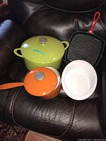 Cookware set on leather chair showing green round pot with lid, orange saucepan with lid, red grill pan, and white ceramic casserole dish.