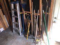 Full view of all items showing the aluminum ladder and various wooden-handled garden tools grouped together in a shed corner.