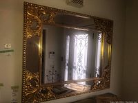 Full view of large rectangular mirror with ornate golden frame showing intricate scrollwork and detailing, reflecting a door with decorative grille.