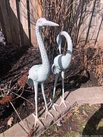Front view of two metal crane sculptures side by side on garden border showing full height and legs