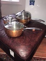 Three copper pots with glass lids and stainless steel handles set on a dark brown leather surface.