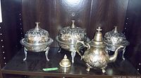 Full set of ornate metal serving pieces including three lidded bowls on legs, one teapot, and one small covered container, arranged on a dark wood shelf.