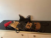 Photo showing 7 purses of various styles and colors laid out on a dark surface against a wall. Includes red, tan, black, and patchwork purses with straps and zippers.