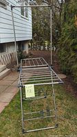Clothes drying rack fully assembled outdoors on grass and sidewalk, showing metal bars and black plastic connectors.
