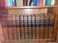 Photo of volumes 1 to 11 of Encyclopedia Britannica 200th Anniversary Edition on wooden shelf, showing leather bindings with gold embossed spine titles and volume numbers.