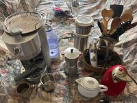 Mix of small kitchen appliances and utensils on patterned fabric including espresso maker, can opener, stovetop coffee pot, teapot, thermal flask, canisters, wooden spoons, knives, and decorative vegetables.